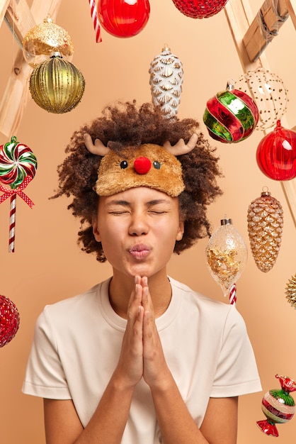 Free photo curly woman stands with closed eyes has pleading expression keeps palms pressed together asks for help says please wants friend to stay and celebrate new year with her poses over decorated background