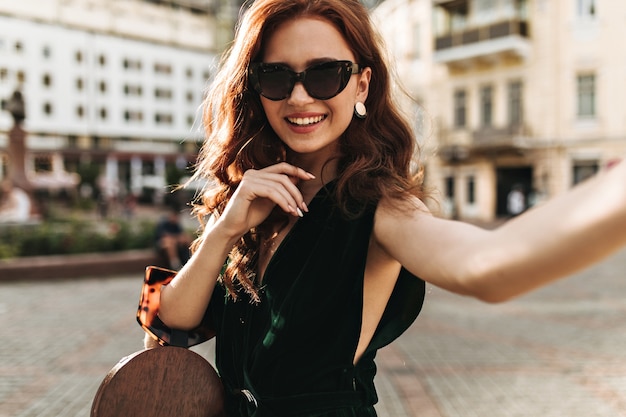 Curly woman in velvet dress holds handbag and takes selfie outside