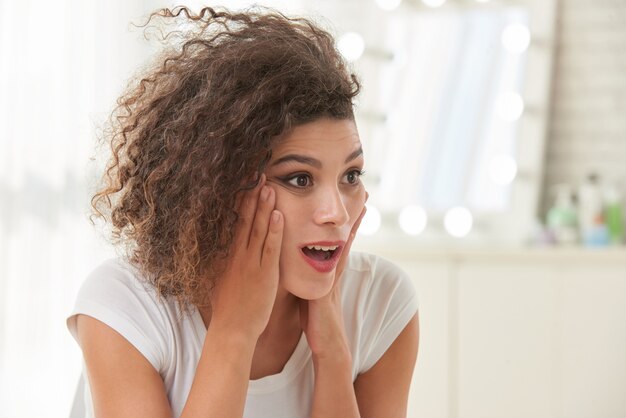 Curly young Caucasian woman posing with surprised face and hands on cheeks