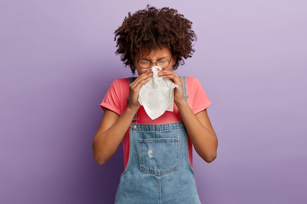 Free photo curly young female feels unwell, blows nose in white tissue, suffers from running nose, cold symptoms or allergy