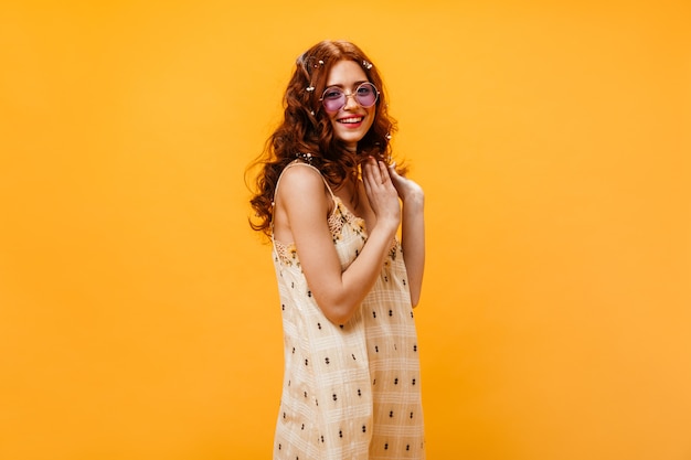 Free photo curly young woman dressed in checkered summer dress and sunglasses posing on orange background.