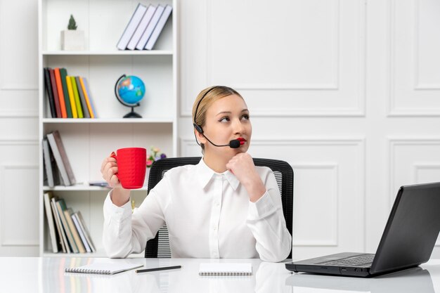 Customer service pretty blonde girl in white shirt with laptop and headset holding a red cup