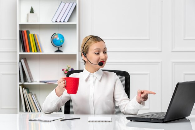 Customer service pretty blonde girl in white shirt with laptop and headset pointing on screen