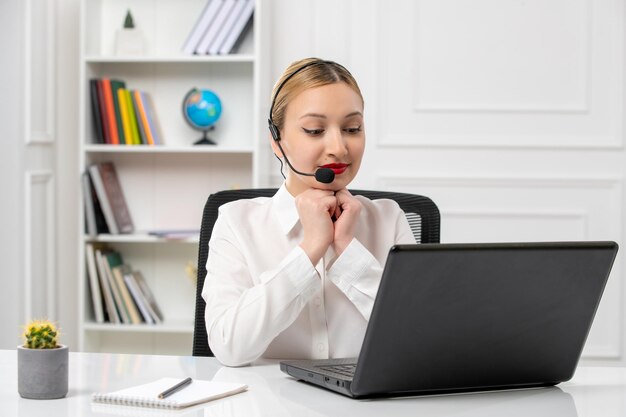 Customer service pretty blonde girl in white shirt with laptop and headset putting hands on chin