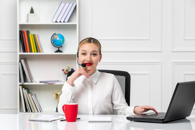 Customer service pretty blonde girl in white shirt with laptop and headset smiling with red cup