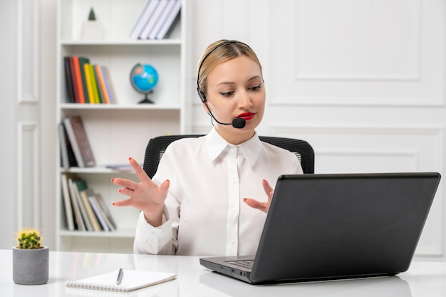 Customer service pretty blonde girl in white shirt with laptop and headset waving hands listening