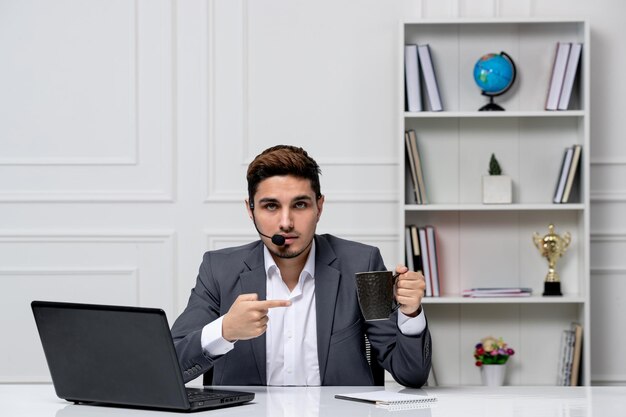 Customer service pretty gentleman with computer in grey office suit pointing at coffee cup