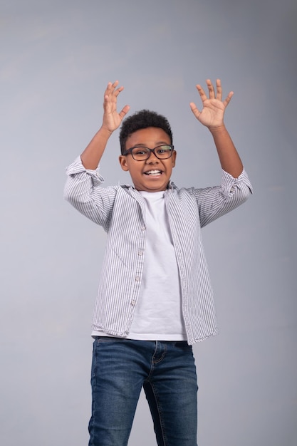 Cute African American boy in trendy eyeglasses standing with his arms raised before the camera