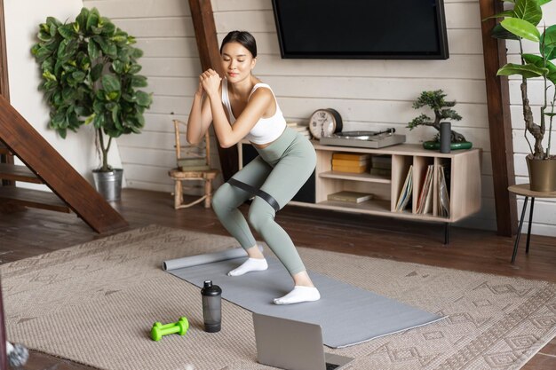 Cute asian fitness girl at home doing workout squats with stretching elastic rope on legs standing o...