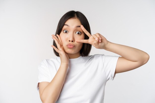 Cute asian girl posing with kawaii vsign peace gesture near face standing in tshirt over white background Copy space