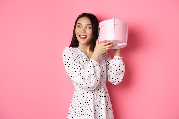 Cute asian woman shaking box with gift smiling and looking intrigued guess what inside present stand...