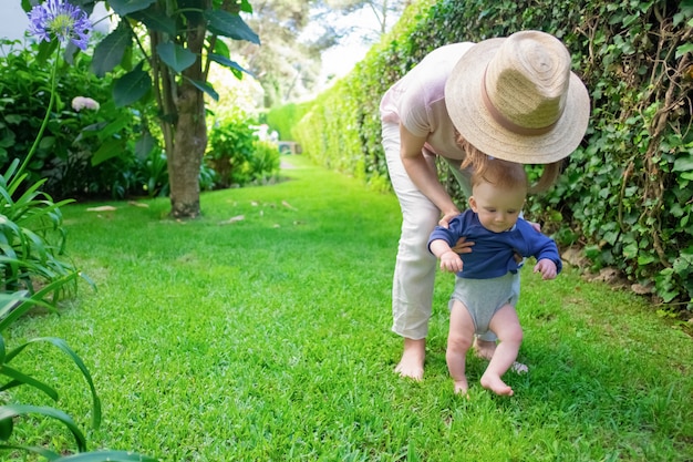 Cute baby in blue shirt doing first steps with help of mom and smiling. Young mother in hat holding infant on grass. First barefoot steps 