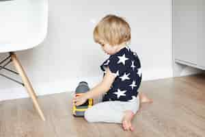 Free photo cute baby boy with blonde hair sitting on wooden floor in his bedroom, holding his favourite toy and smiling. toddler having fun, playing with yellow plastic truck. early learning.