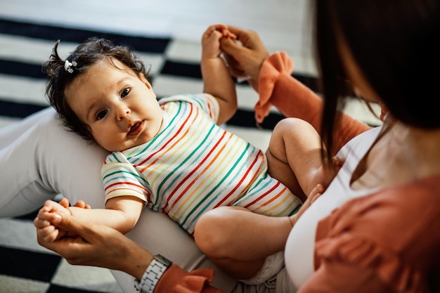 Cute baby girl lying in mother's lap while holding her hands and looking at camera