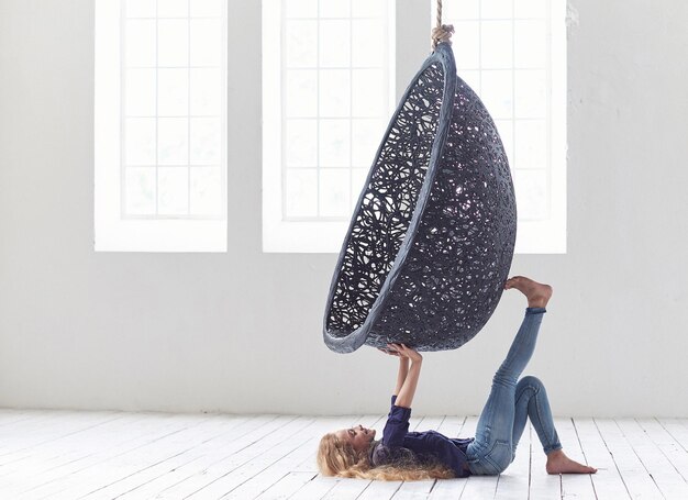 A cute beautiful little girl in casual clothes laying under a suspended chair in an empty studio.