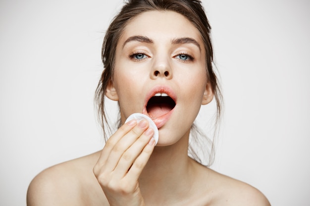 Cute beautiful natural brunette girl cleaning face with cotton sponge smiling looking at camera over white background. Cosmetology and spa.