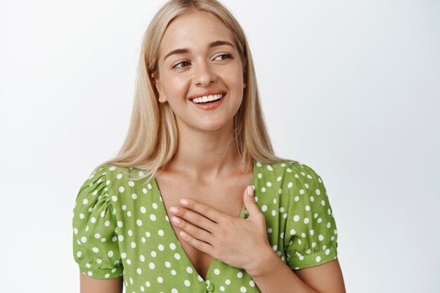 Cute blond girl in dress, holding hand on chest, smiling and looking aside on white. Copy space