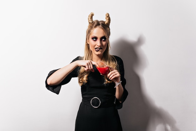 Free photo cute blonde vampire in dress standing on white wall. pretty slim girl drinking blood in halloween.