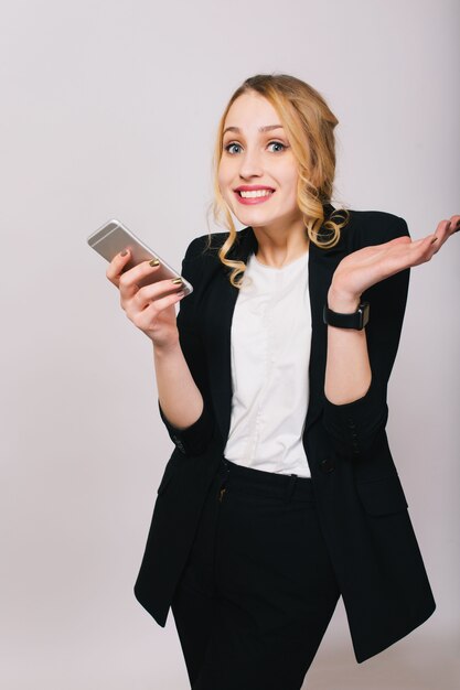 Cute blonde young office woman in white shirt, black suit with phone looking isolated. Expressing true positive emotions, success, work, friendly