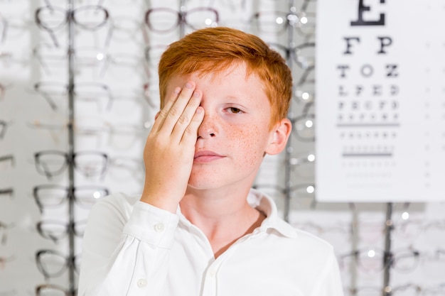 Free Photo cute boy covered his eye with hand standing in optics clinic