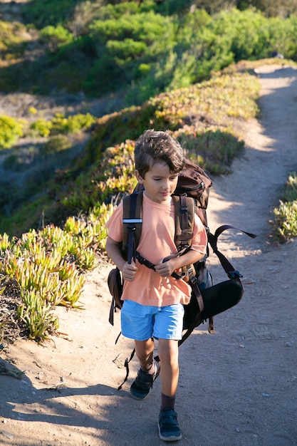 Free Photo cute boy walking on countryside path and carrying huge backpack. front view, full length. childhood or adventure travel concept