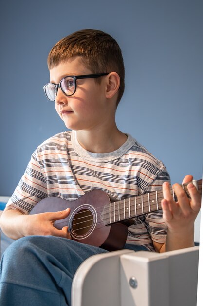 A cute boy with glasses learns to play the ukulele guitar at home, in his room.