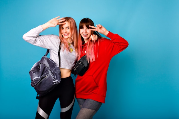 Cute bright portrait of two happy pretty hipster girls wearing sportive clothes for fitness and backpack, smiling and hugs, blue wall.