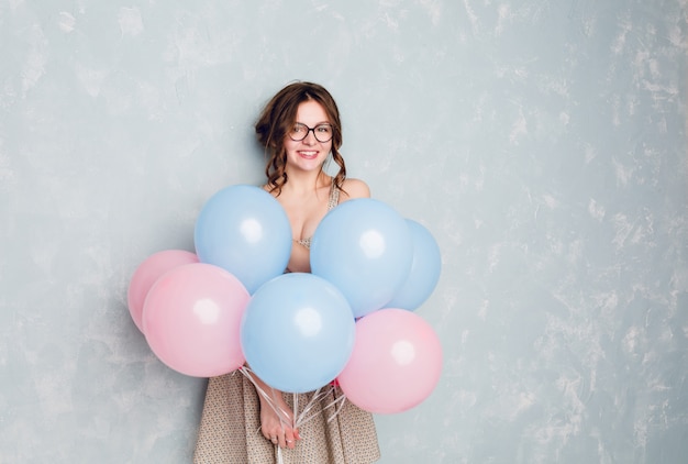 Free photo cute brunette girl standing in a studio, smiling widely and holding blue and pink balloons.