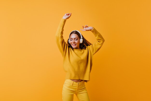 Cute brunette lady in yellow pants enjoying good song.  carefree woman in soft sweater dancing on orange wall.