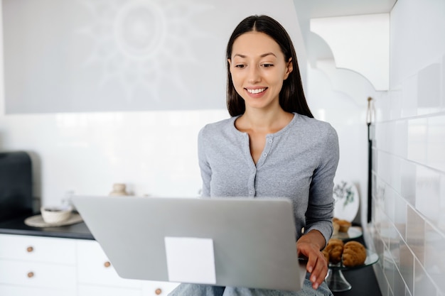 Cute brunette woman sitting on the table at kitchen and using laptop