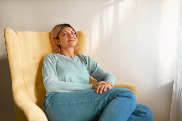 Cute carefree young Caucasian female wearing casual clothes sitting in yellow armchair on white curtain