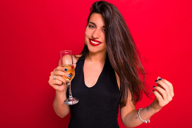 Cute caucasian elegant woman with red lips and long dark hair playfully looking to camera and drinking champagne.