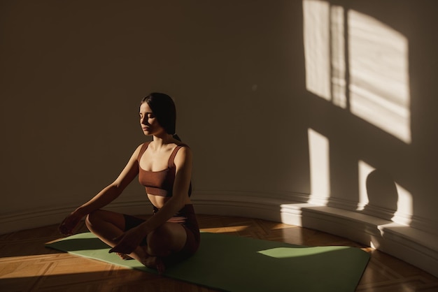 Free photo cute caucasian young woman meditates while sitting on mat near light wall space for text darkhaired girl wears top and leggings in training zen concept