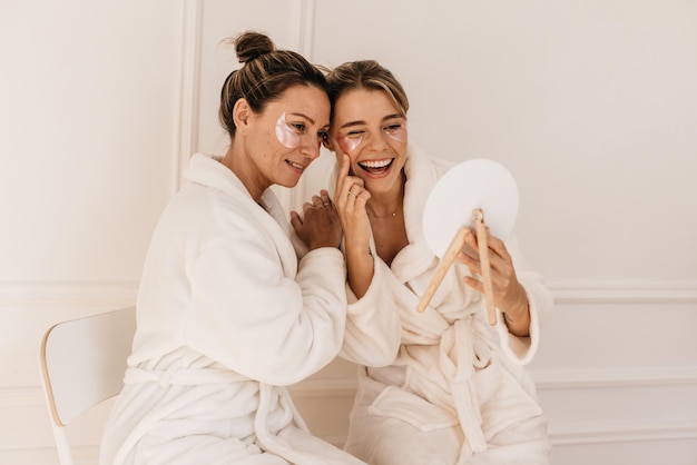 Free photo cute caucasian young women in bathrobes admire themselves while looking in mirror on white background. beauty and personal care, skin and face care concept