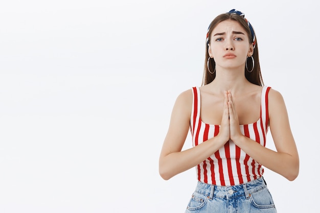 Free photo cute and charming young woman in striped top and headband pursing lip frowning and holding hands in pray while apologizing or being in need and asking for help