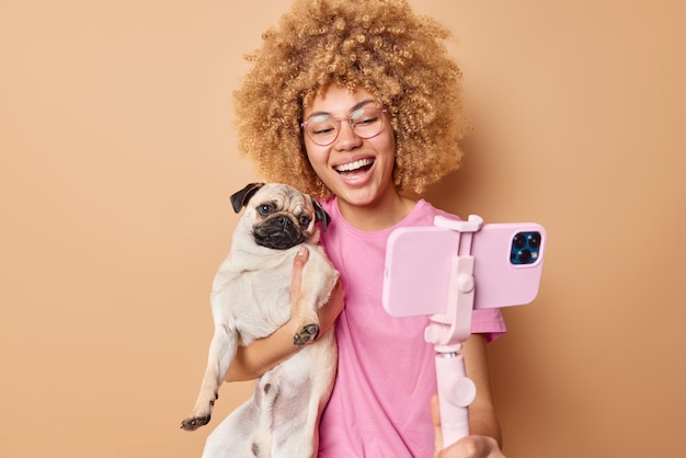 Free photo cute cheerful female pet owner poses with pug dog for selfie spend free time together make memorable photo while having walk isolated over brown background glad young woman with domestic animal