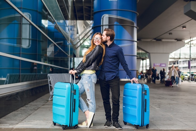 Cute couple with suitcases is standing outside in airport. She has long hair, glasses,  yellow sweater, jacket. He wears black shirt, beard. Guy is hugging and kissing the girl.