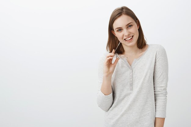 Cute coworker have and eye on charming guy flirting during break biting eyeglasses holding eyewear in hand smiling tenderly posing against gray wall in casual white blouse