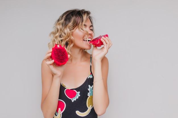 Free photo cute curly girl eating pitaya with pleasure. indoor photo of fair-haired caucasian woman enjoying exotic fruit.