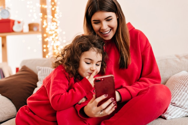 Cute curly girl using smartphone with mother. Smiling young mom chilling with preteen daughter on sofa.