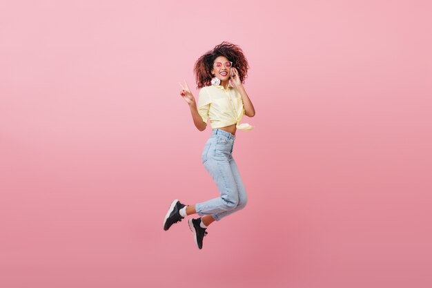 Cute curly woman in yellow shirt expressing happy emotions with smile. Magnificent african hipster girl jumping.