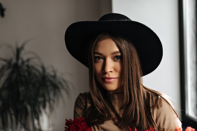 Free photo cute dark-haired woman with tanned skin looks into camera. woman in black hat and beige t-shirt is posing in bright room.