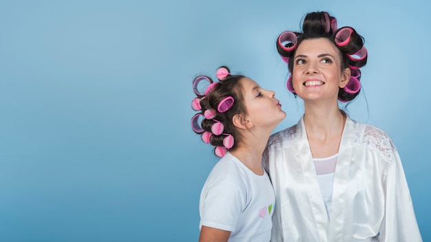 Cute daughter kissing mother in curlers and bathrobe 