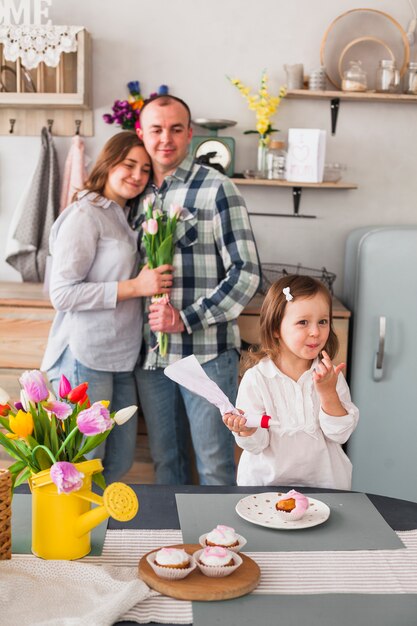 Cute daughter making cupcake near parents with flowers