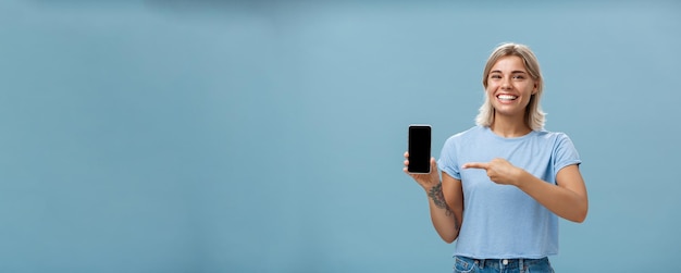 Free photo cute enrgized girl showing smartphone charming blonde young female with cool tattoos laughing and smiling from happiness and joy pointing at device holding phone with screen faced to camera