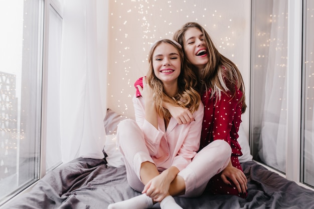 Cute european girl in pink sleepwear posing with charming smile in bedroom. Indoor portrait of fascinating ladies in pajamas hugging and laughing.