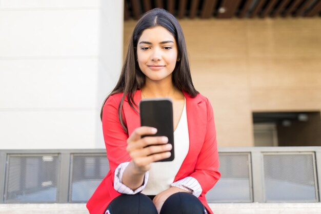 Cute female executive reading incoming notification on smartphone while sitting outside office