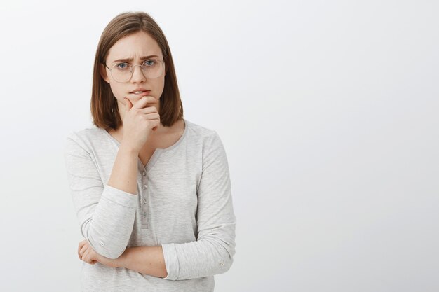 Cute female geek trying solve hard math riddle standing thoughtful over white wall rubbing chin during brainstorm looking while making decision or thinking posing over gray wall