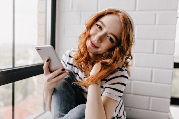 Cute female model in headphones looking with smile. Appealing ginger woman with phone in hand sitting at home.