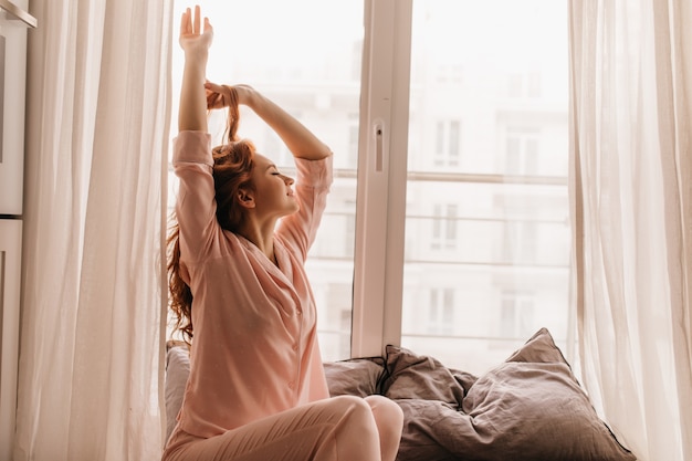 Free photo cute female model in pink pajama enjoying morning. pleasant ginger woman sitting on bed.